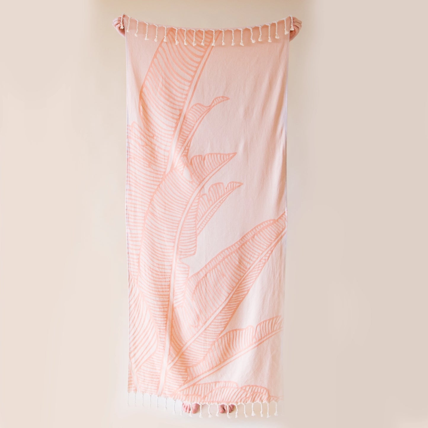 On a beige background is a model holding up the Bird of Paradise Beach Towel with the bird of paradise leaf print and string tassels on each end.