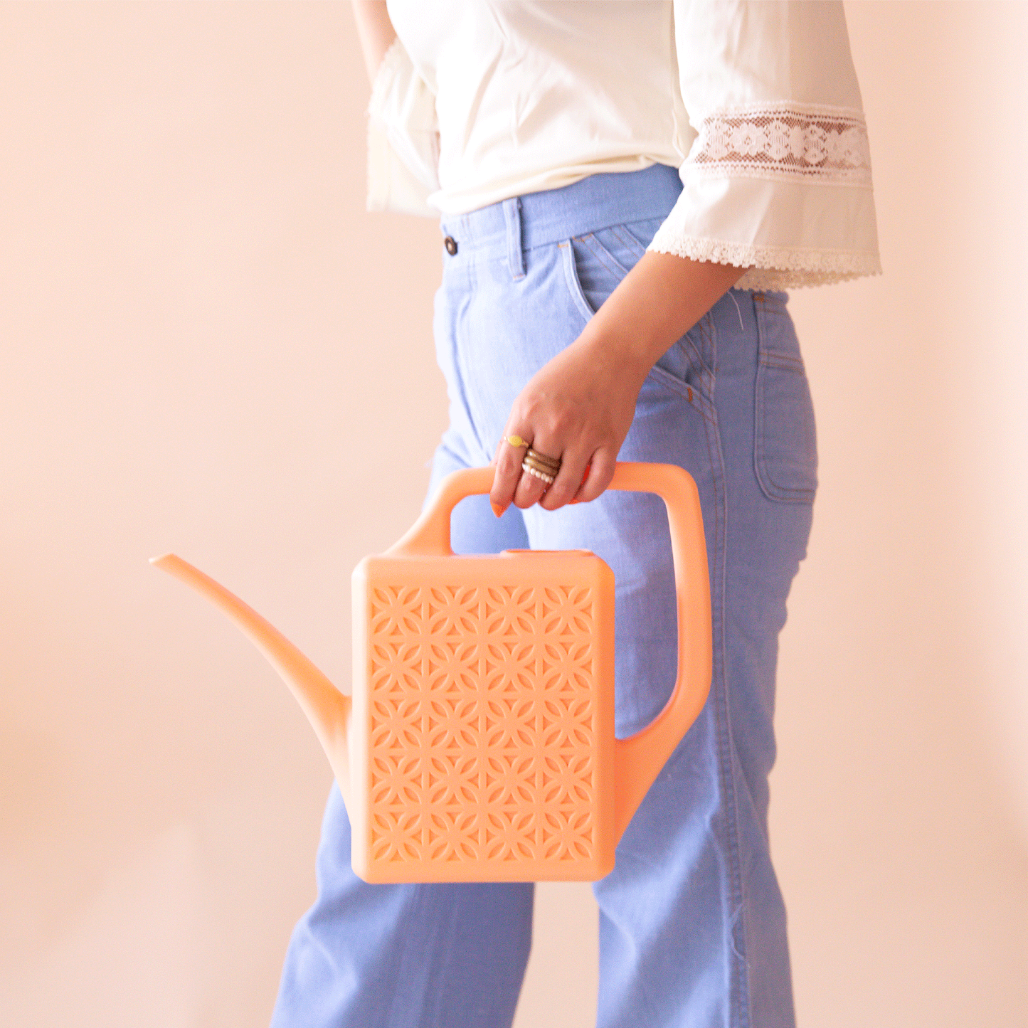 On a peach background is a model holding the breeze block watering can in the shade peach.
