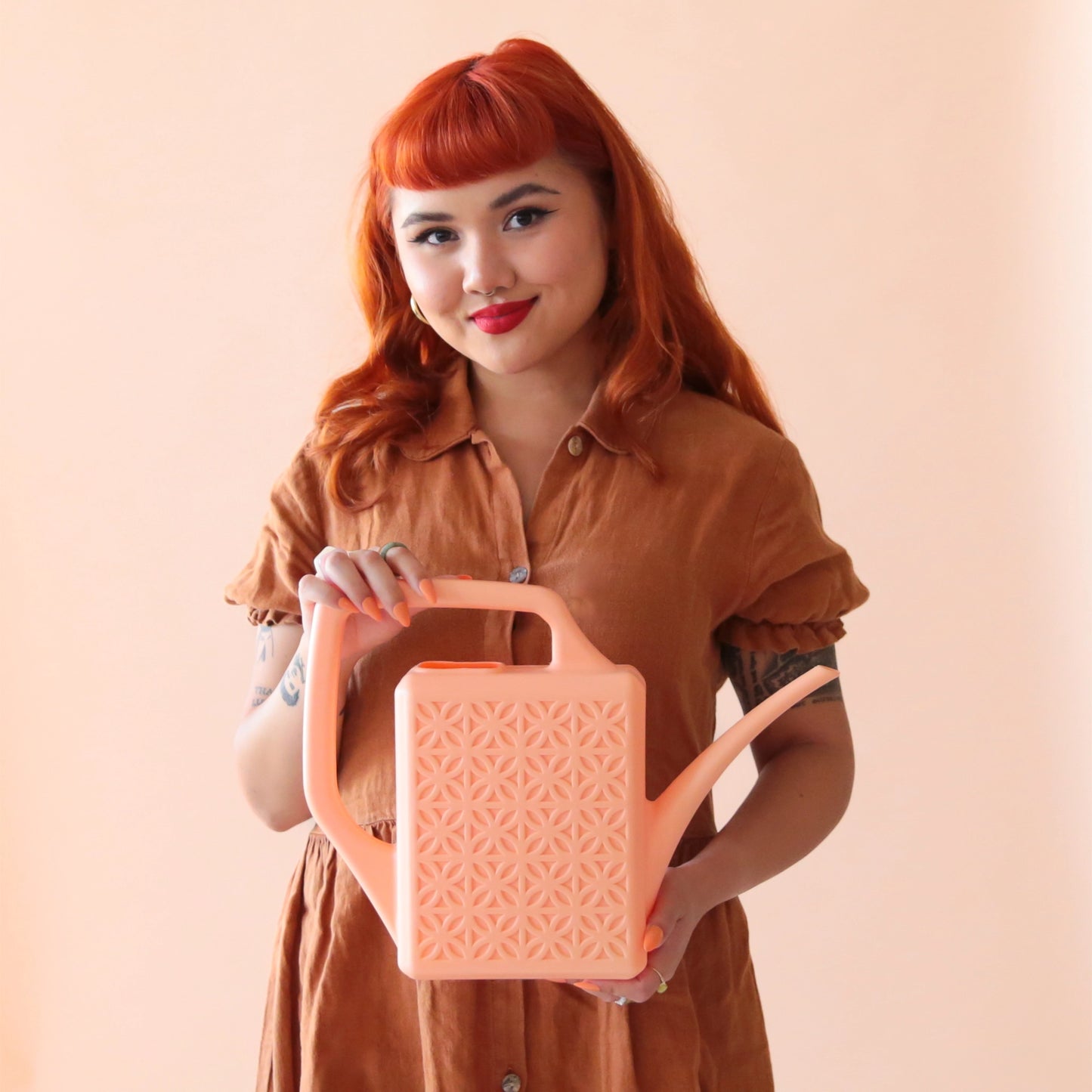 On a peach background is a model holding the breeze block watering can in the shade peach.