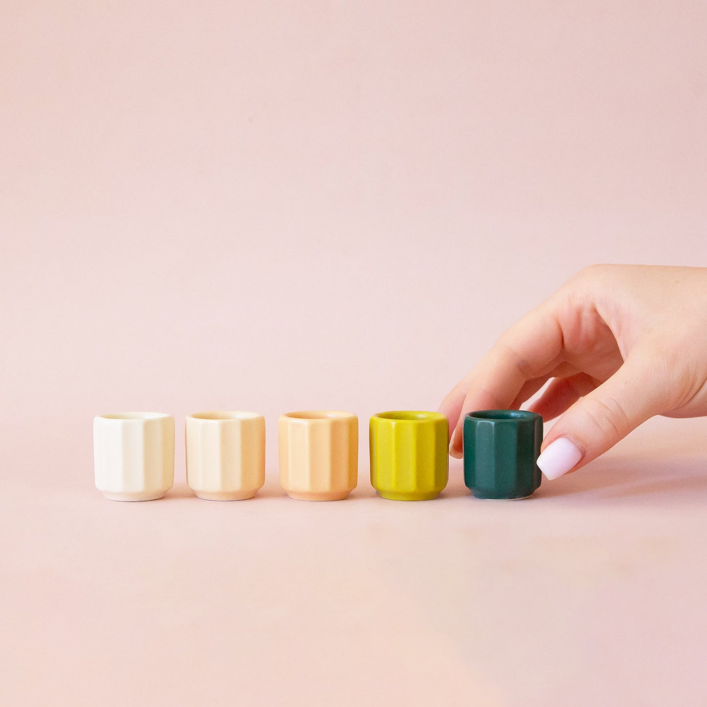 A hand comes into frame on the right hand side of the image. The hand reaches for a miniature aquamarine colored pot. The pot is last in a row of 5 tiny pots. They go in order from white to ivory to peach to chartreuse to the final aquamarine color. The background of the image is a soft pink color.