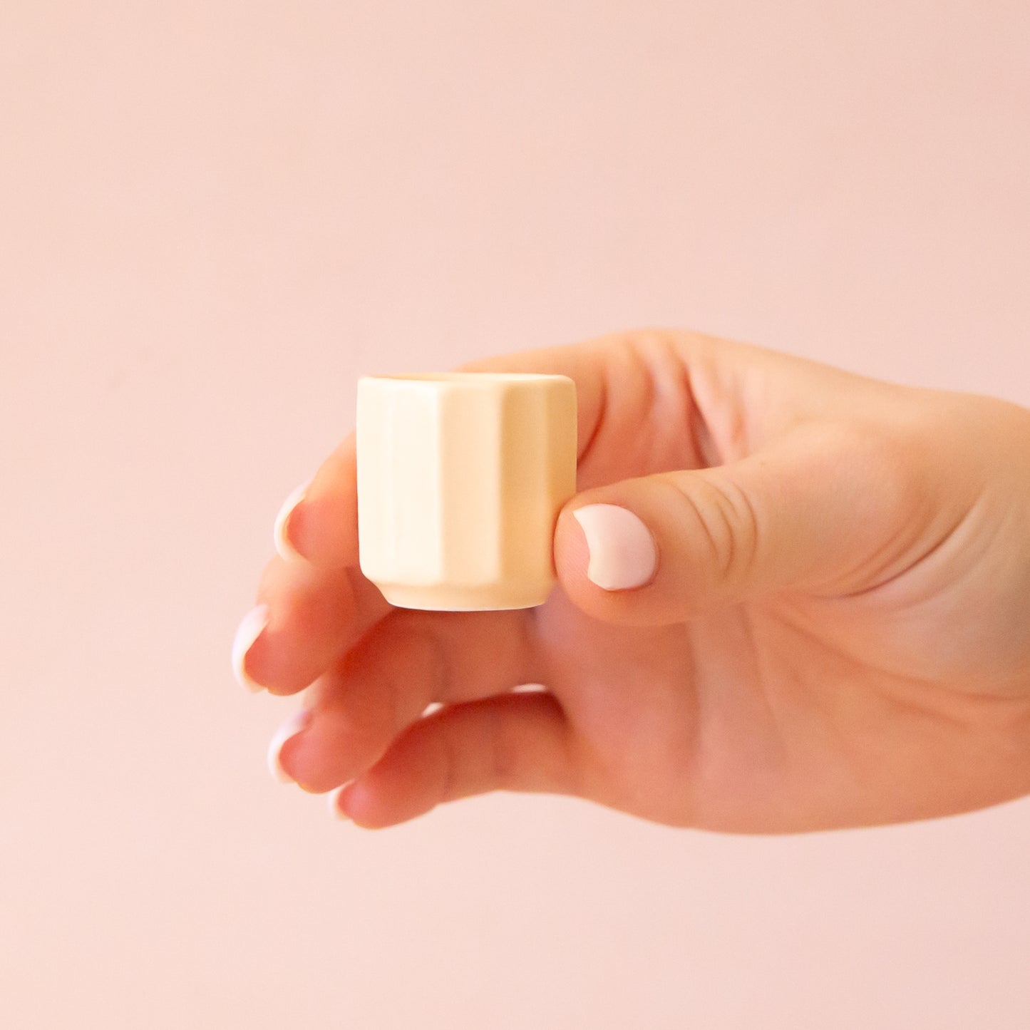 A hand comes into frame on the right hand side of image with soft pink nails. The hand is holding an itty bitty pot about the size of thimble that is a soft tan color. The background is all a soft white color.