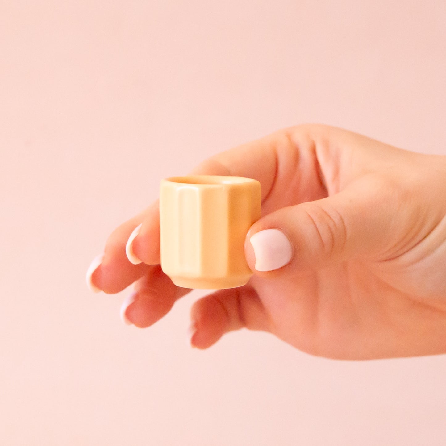 A hand comes into frame on the right hand side of image with soft pink nails. The hand is holding an itty bitty pot about the size of thimble that is a soft peach color. The background is all a soft white color.