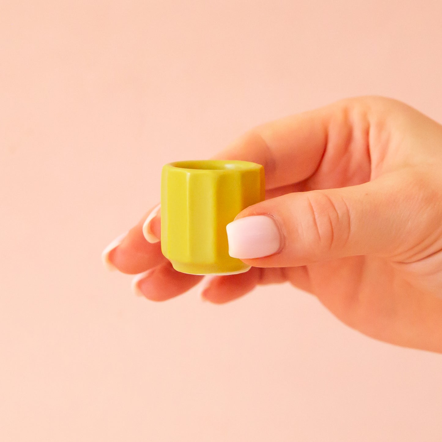 A hand comes into frame on the right hand side of image with soft pink nails. The hand is holding an itty bitty pot about the size of thimble that is a chartreuse color. The background is all a soft white color.