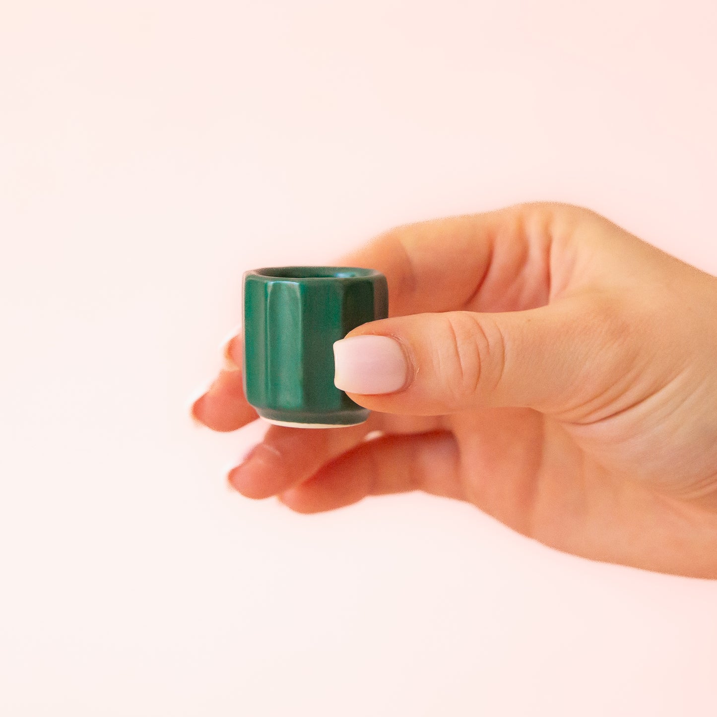 A hand comes into frame on the right hand side of image with soft pink nails. The hand is holding an itty bitty pot about the size of thimble that is a dark blue green color. The background is all a soft white color.