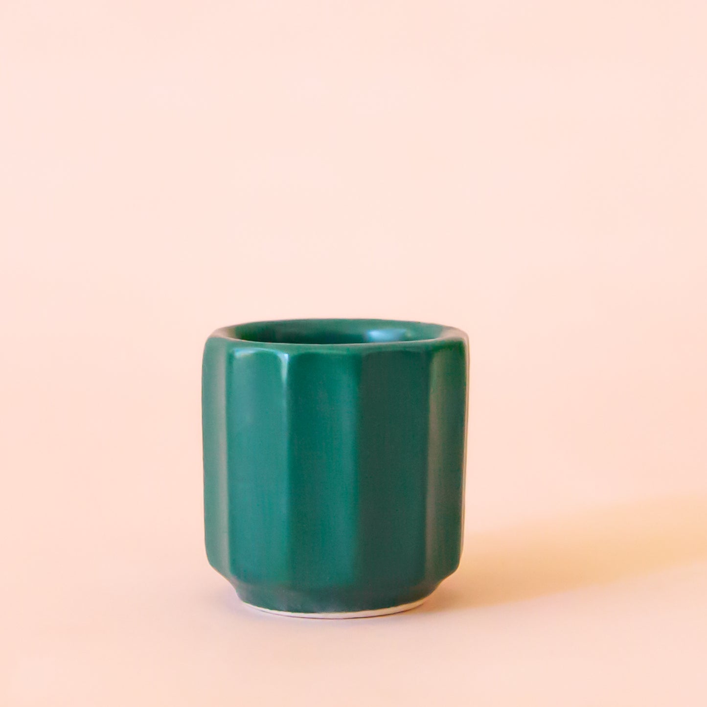 An itty bitty pot about the size of thimble that is a dark blue green color sits in the middle of the image. The pot hat little ridges running around it. The background of the image is a soft white color.