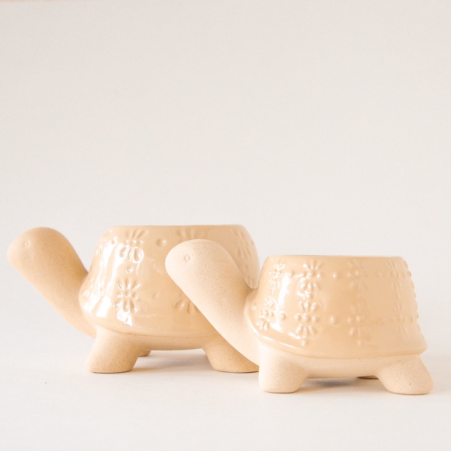 On a white background is two different sized ceramic planters in the shape of a turtle and has a light tan "shell" with a subtle floral texture.