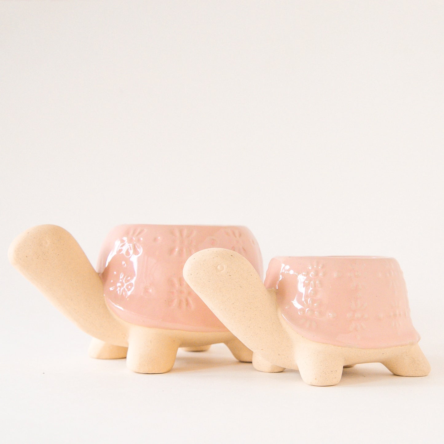 On a light grey background is two different sized ceramic planters in the shape of a turtle with a light pink "shell" with a subtle floral print.