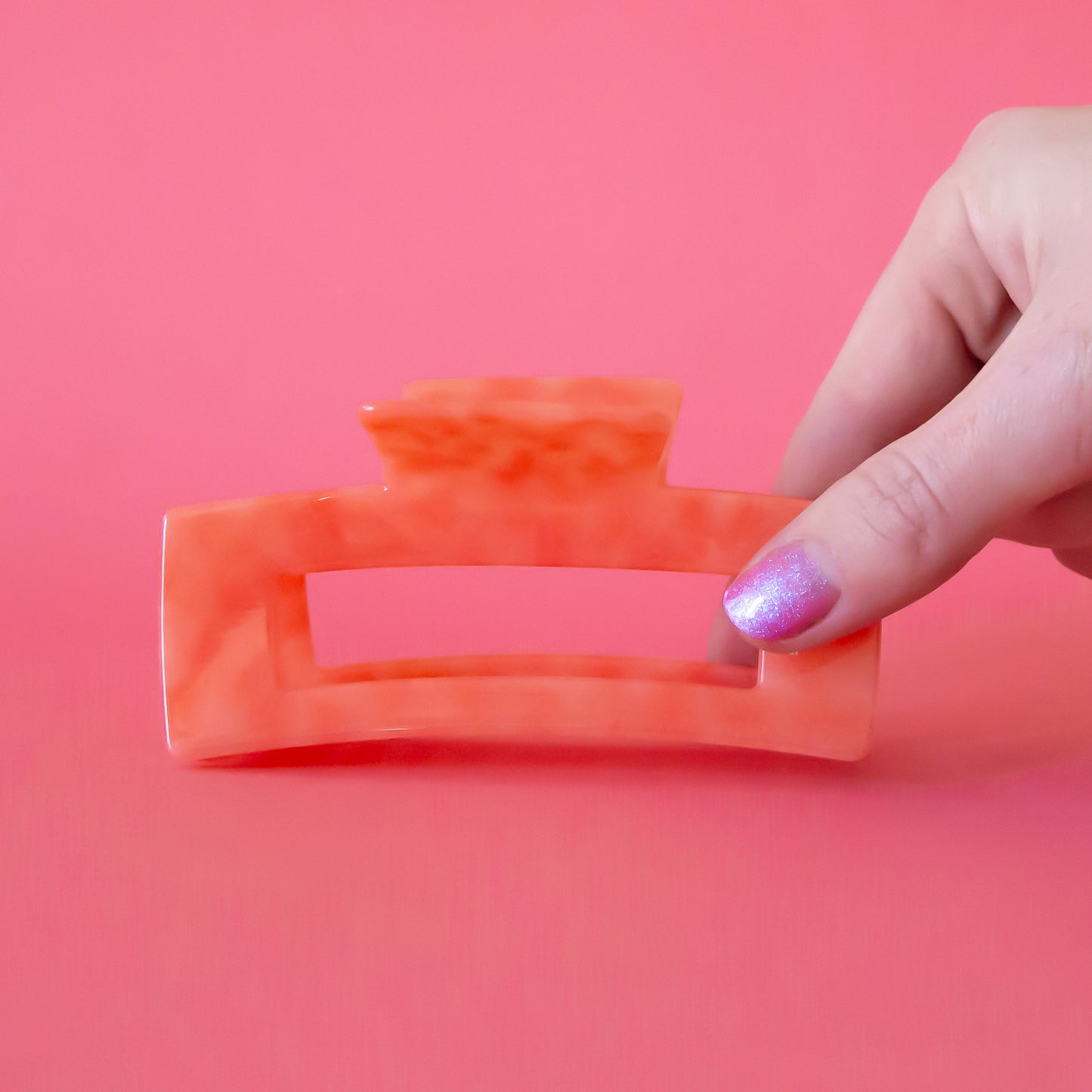 A beautiful coral colored hair claw sits in the middle of page. A hand reaches into frame from the right hand side holding onto the hair claw. The thumb nail is painted with glitter. The background is a watermelon color.