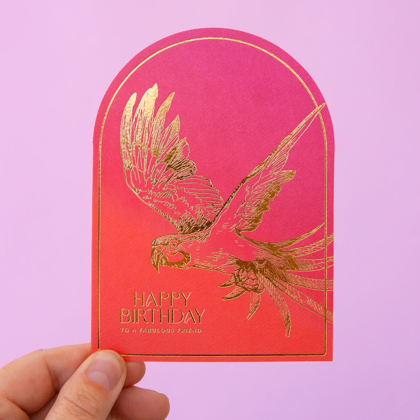 A hot pink and orange ombre colored arched shaped card with gold foiled text that reads, "Happy Birthday To A Fabulous Friend" along with a gold foiled parrot illustration.