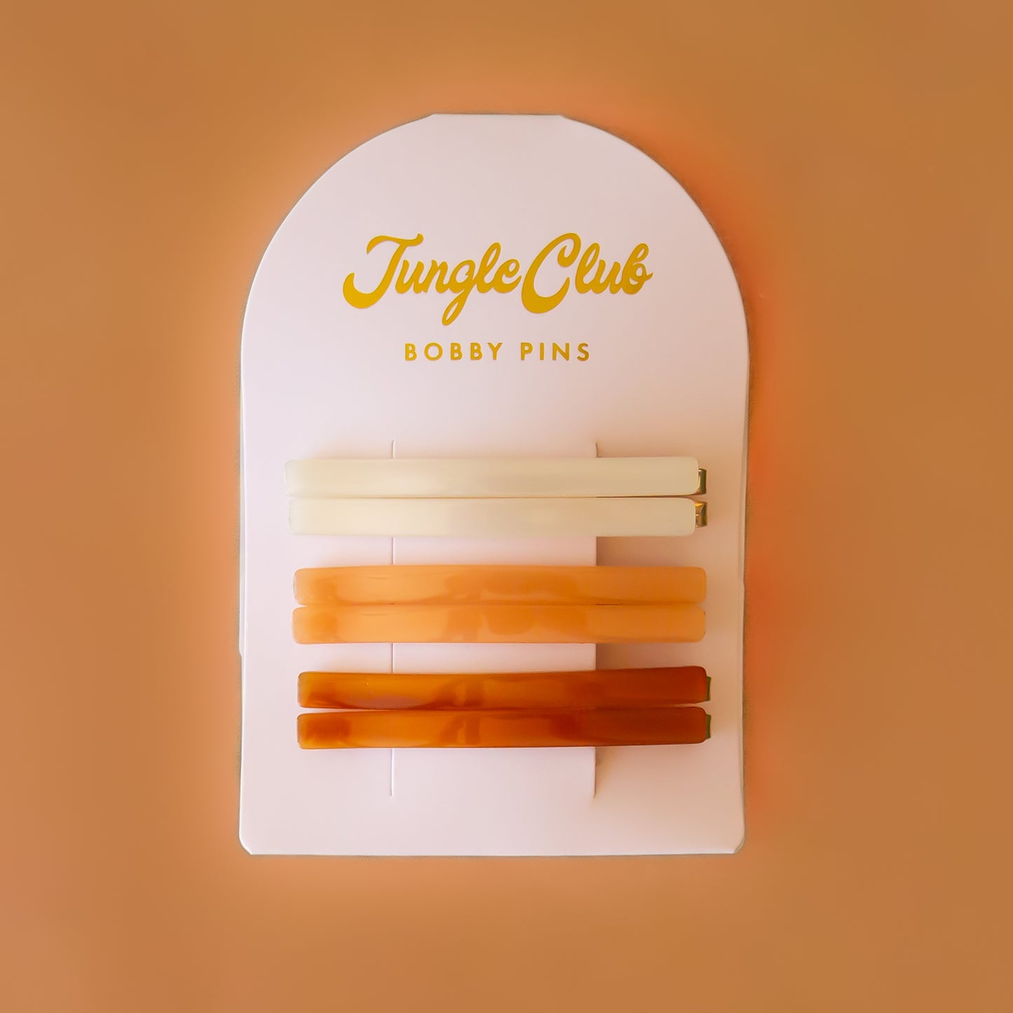 On a pink background is an arched packaging with embossed text at the top that reads, "Jungle Club Bobby Pins" along with six thin hair clips in three colors from an ivory shade to orange to a honey rust color.