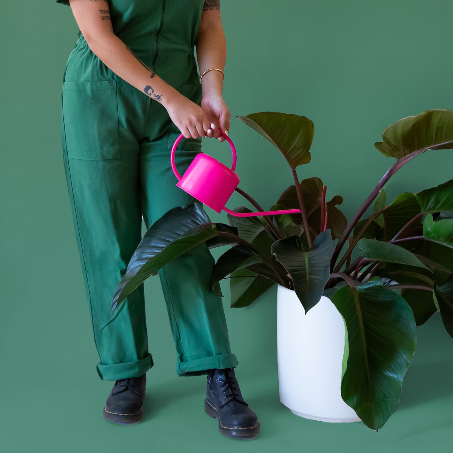 A hot pink metal watering can with a rounded handle and a long spout is held in hands pouring into a large leafy plant. The person is wearing a green jumpsuit and the background of the image is all green.