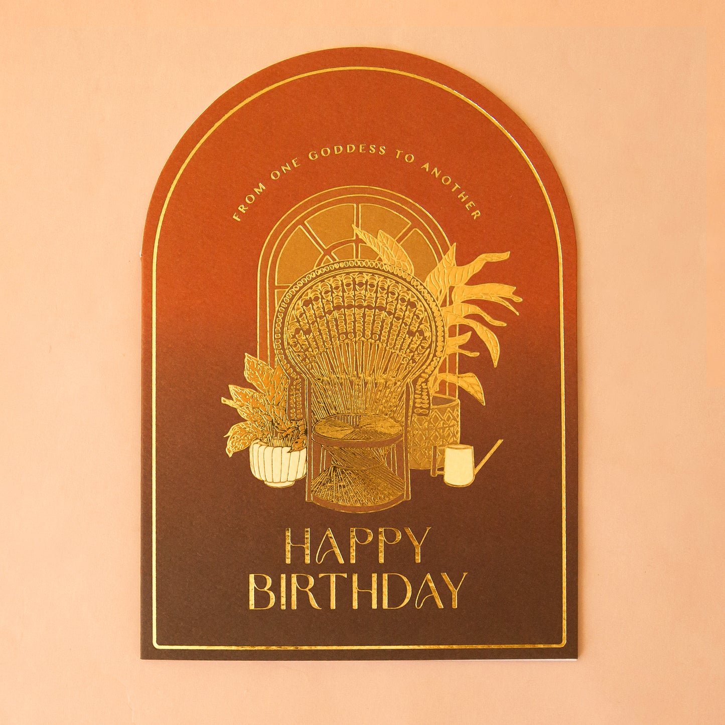 An arched card with orange and brown hues with gold foiled text that reads, 'From One Goddess To Another Happy Birthday' and a design of a peacock chair with plants behind it.