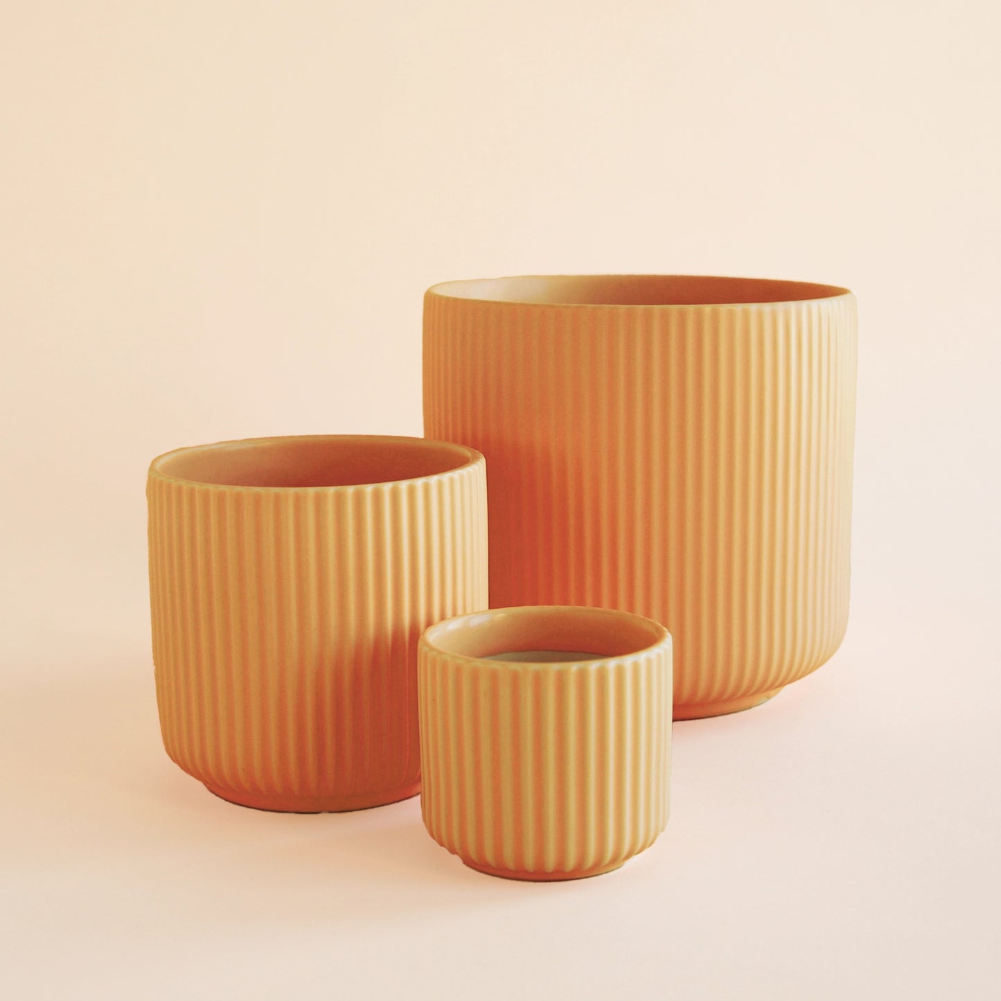 On an ivory background is three different sized orange ceramic planters with a fluted detail and a rounded base.