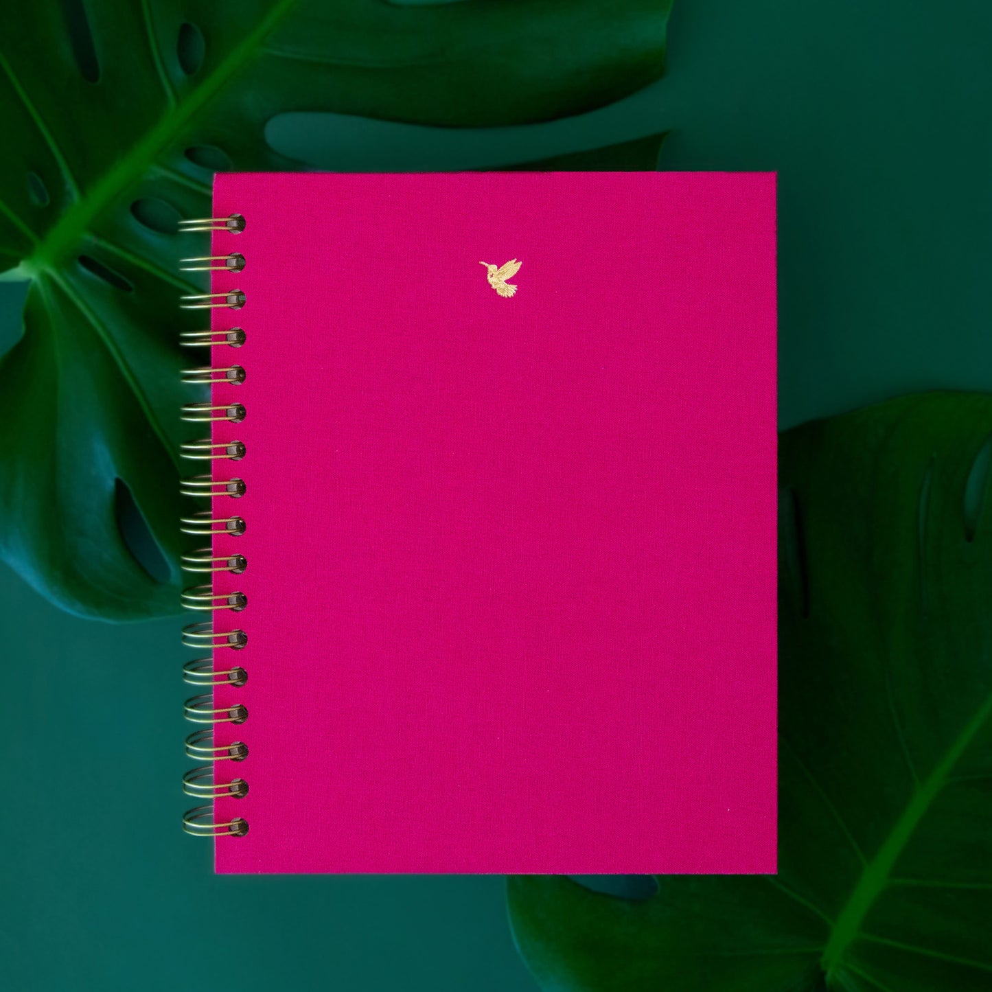 A vibrant pink journal sits on a emerald green background. The journal has a gold hummingbird motif on it.