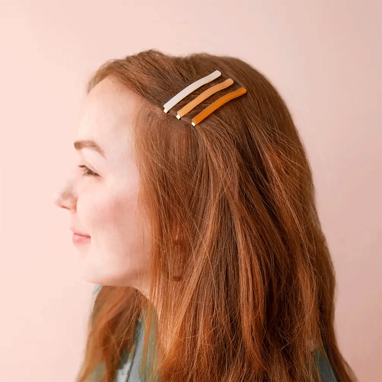 On a pink background is a model wearing all three colors of the thin bobby pin hair clips in her hair.