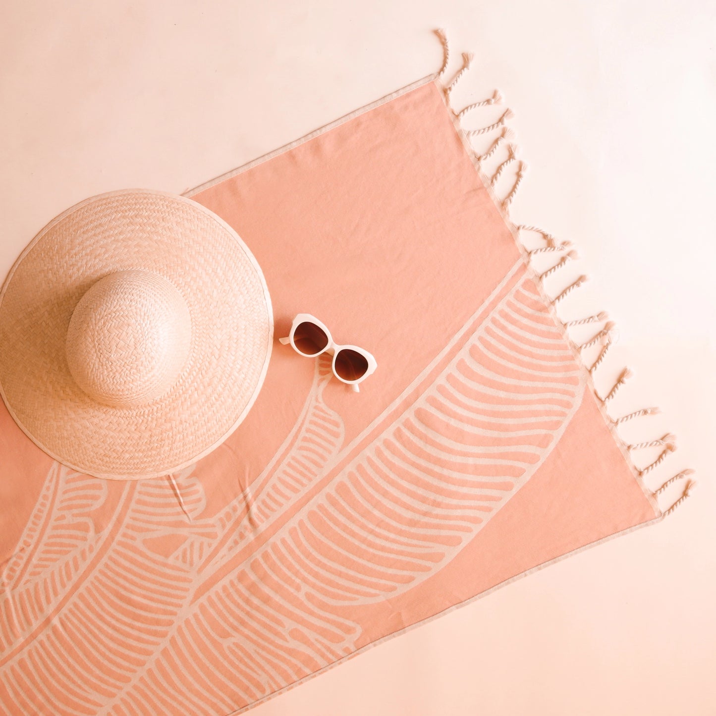 On a light pink background is a salmon colored beach towel with string tassels at each end along with a Bird of Paradise line design.