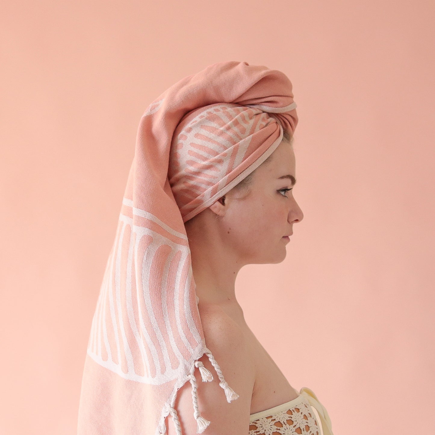 On a pink background is a model holding up the Bird of Paradise Beach Towel with the bird of paradise leaf print and string tassels on each end.