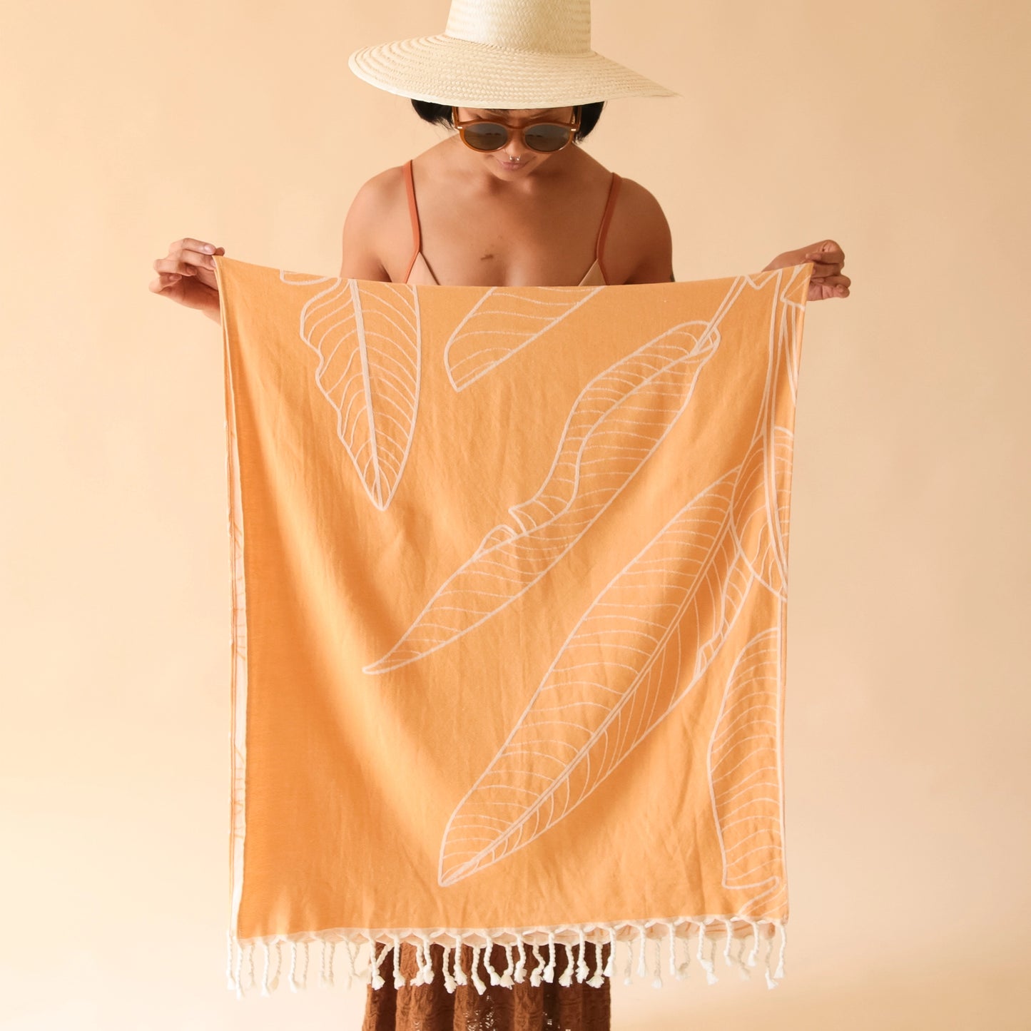 On a tan background is an orange beach towel with a banana leaf print and white tassels on each end.