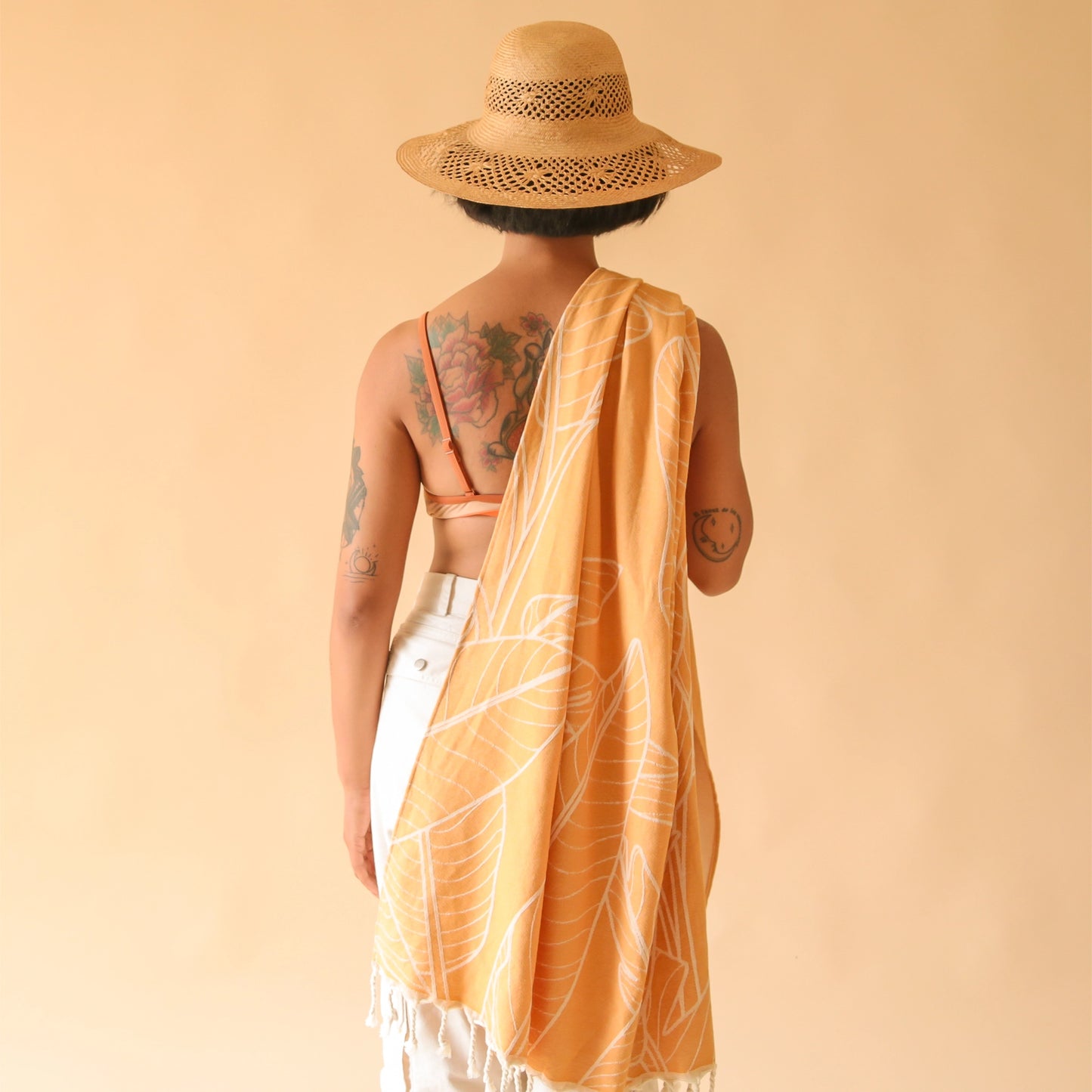 An orange beach towel with a banana leaf print and white tassels on each end.