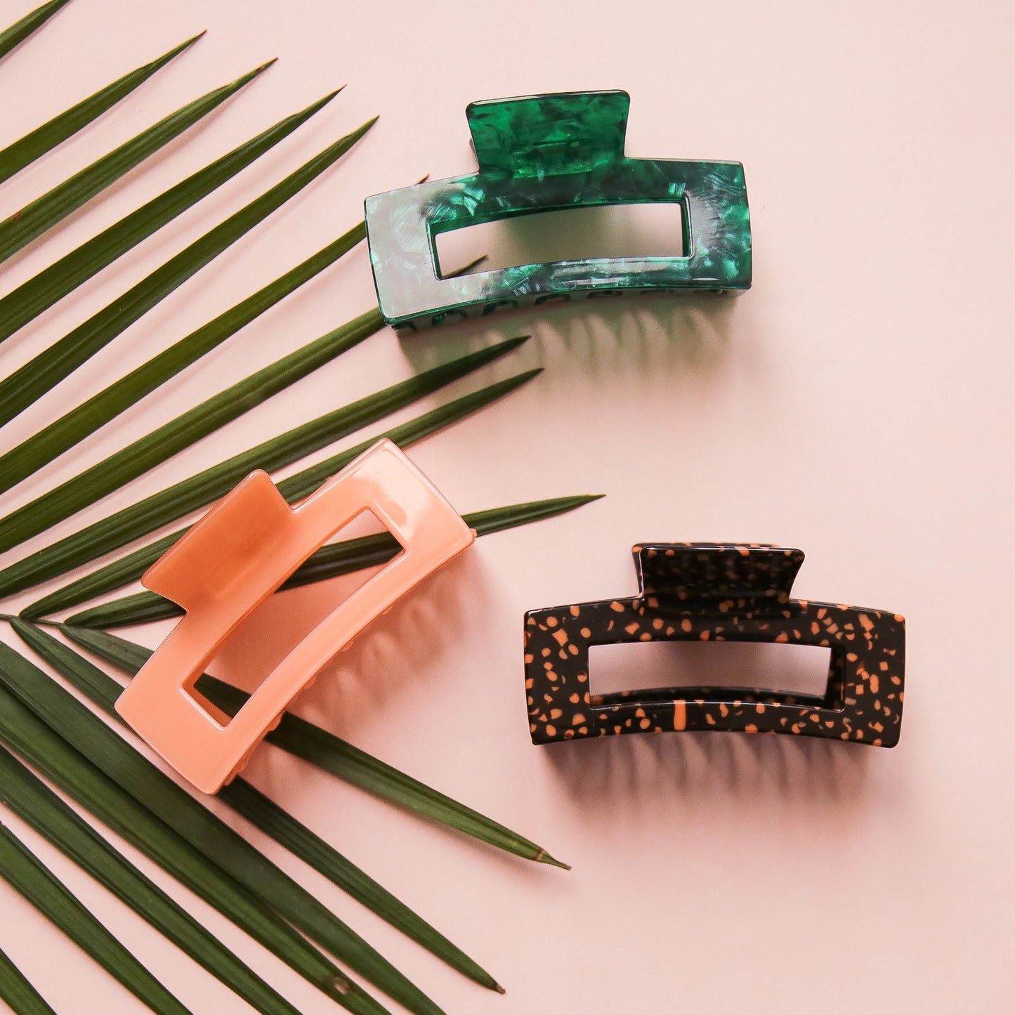On a light pink background is the three different colors available for the Paradise Hair Claw. There is an orange and dark brown leopard print, a dark green shade, and a peachy pink option.