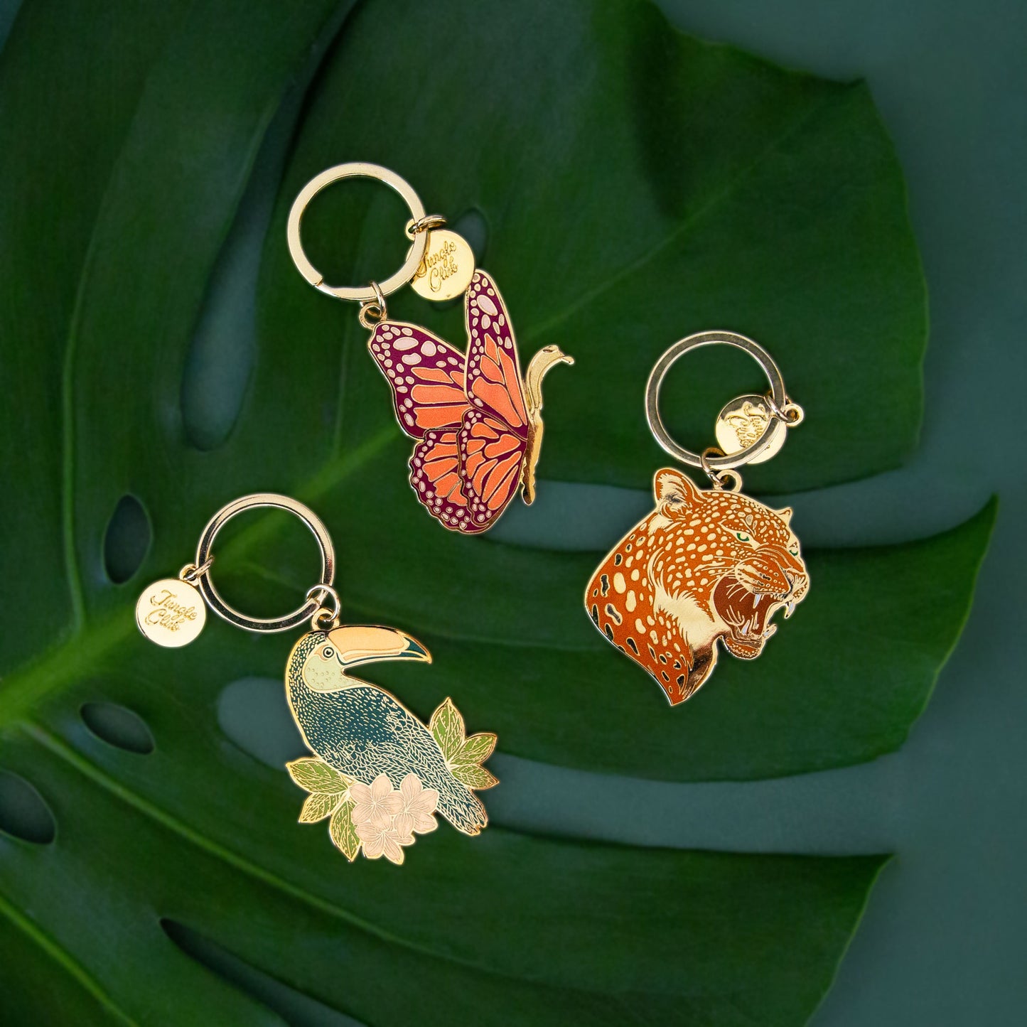 Aerial photo of 3 keychains. One is a butterfly, the second is a toucan and the third is a leopard. The keychains are bright enamel colors and lay on top of a monstera leaf.
