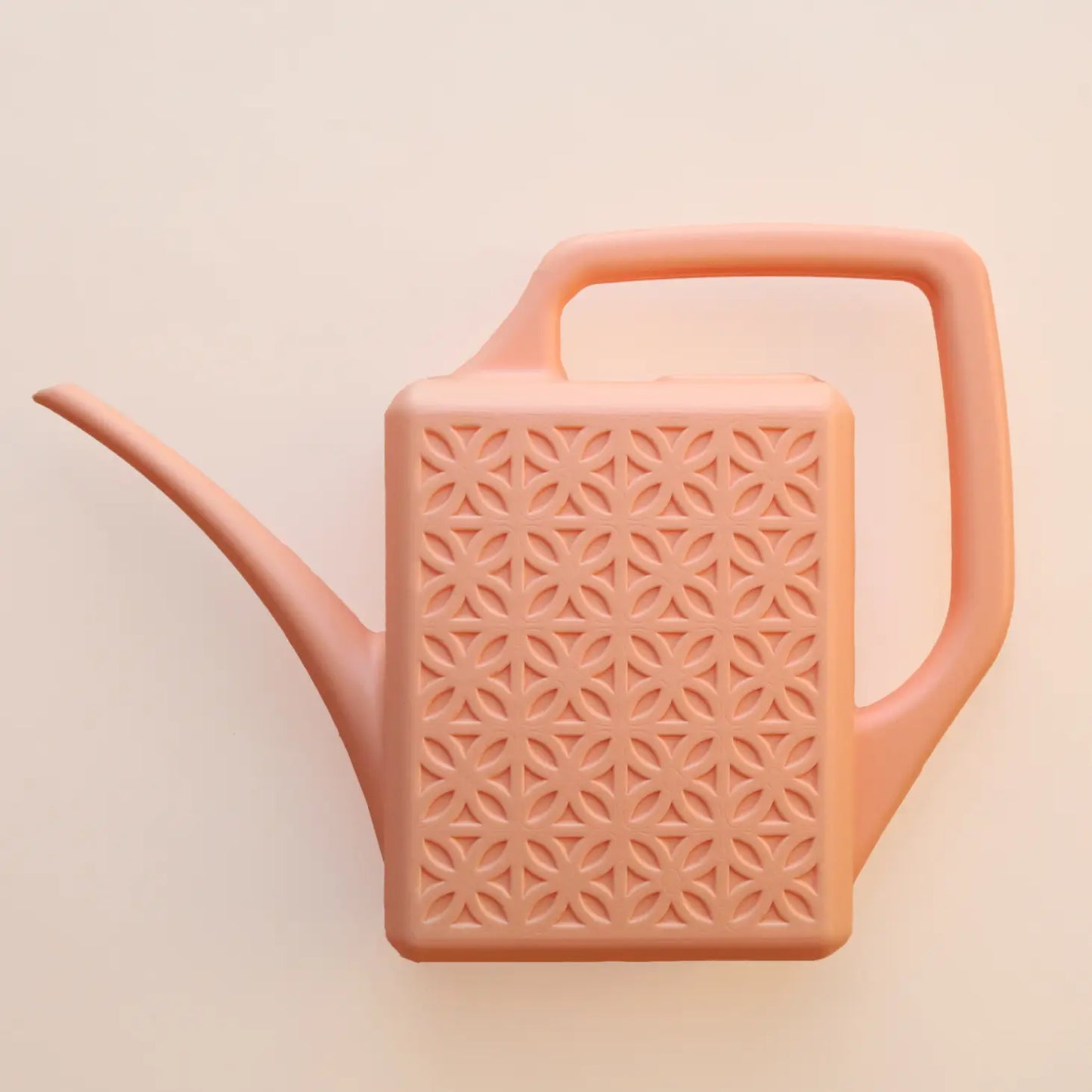 On a peachy background is a light pink plastic watering can with a breeze block design on both sides along with a long spout and a squared off handle for easy watering.