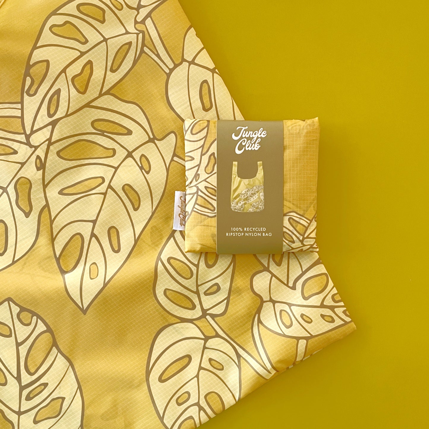 Photographed in front of a green background is a green nylon reusable bag with a lighter green Swiss cheese plant print on the front and back folded in its square condensed pouch.