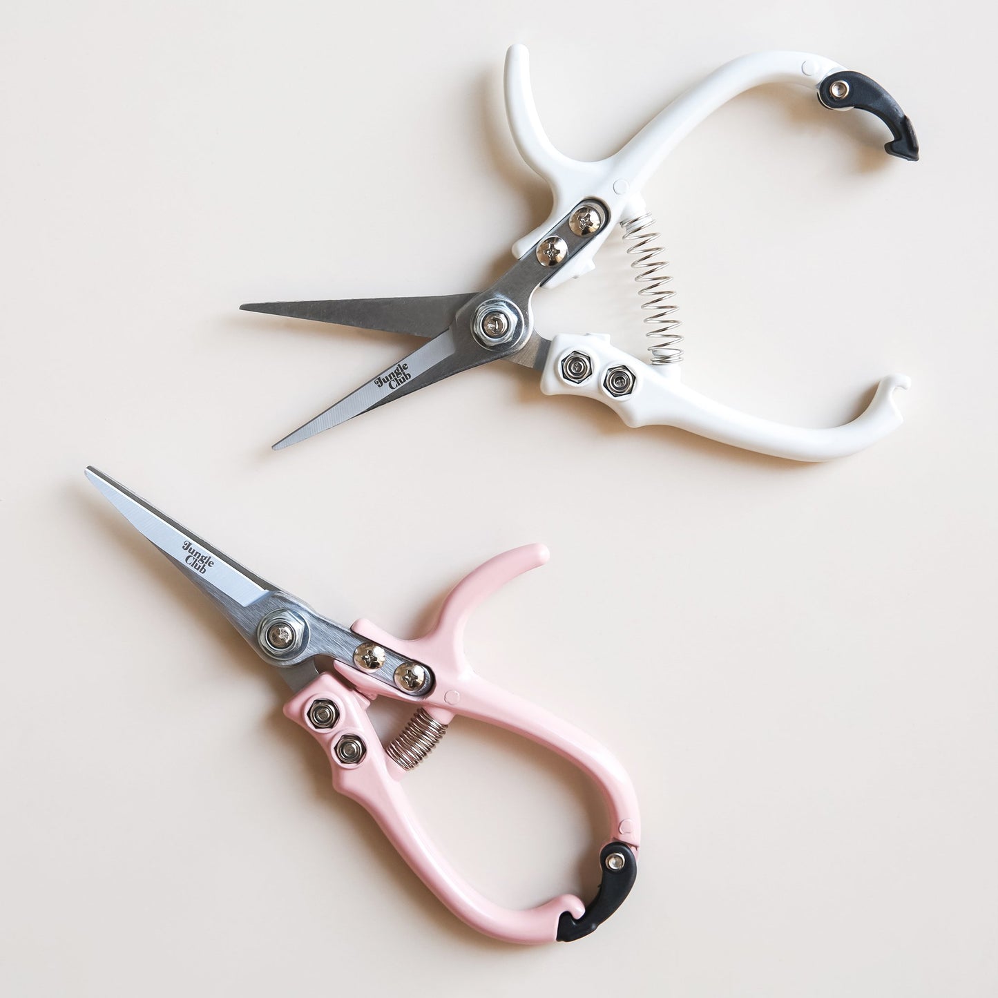 Two pairs of pruning shears laying next to each other. The pair towards the bottom is fasten closed with pastel pink handles and the pair above is positioned unclasped with white handles.