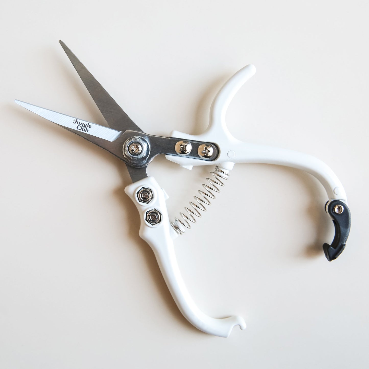 On a cream background is a white pair of pruning shears with silver metal and fixtures and small text on the metal part of the shears that reads, "Jungle Club" in small black letters. In this photograph the clip that keeps the shears closed is unhooked and they are sprung open.