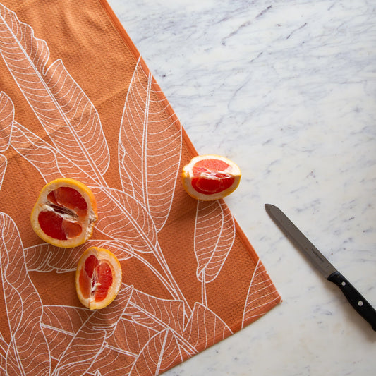 On a marble background is an orange kitchen towel with a white banana leaf design.