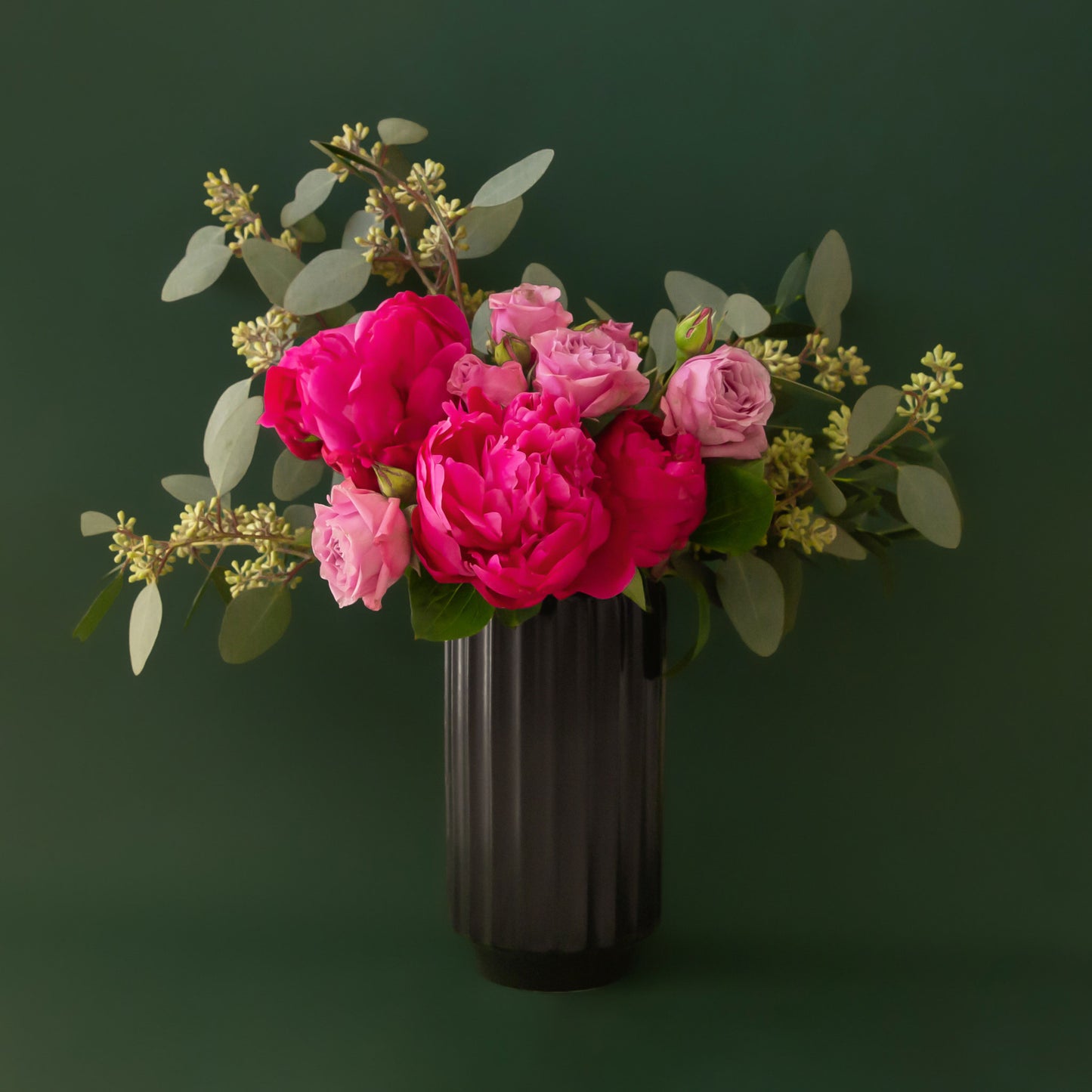 A black fluted vase with pink and lavender roses and peonies and green filler florals spilling out on a dark green background.