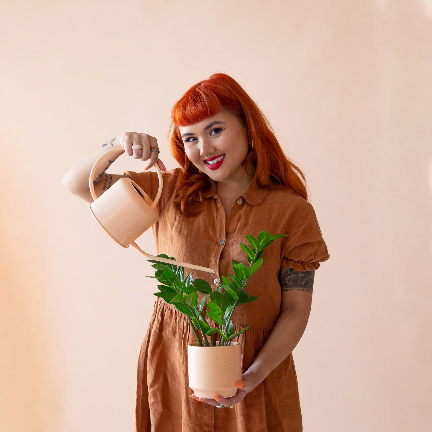 On a peach background is a model holding a house plant and watering with the Cascade Metal Watering Can in the shade Vanilla.
