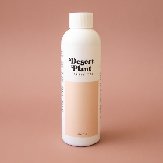 White twist top bottle labeled 'Desert Plant Fertilizer' in black playful lettering. Below the label is a rectangular block of soft beige. The fertilizer sits in front of a solid sand colored background.