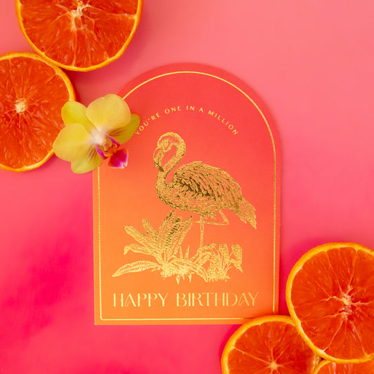 An orange gradient arched shaped card with gold foiled illustrations of a flamingo and text that reads, "You're One In A Million Happy Birthday". 