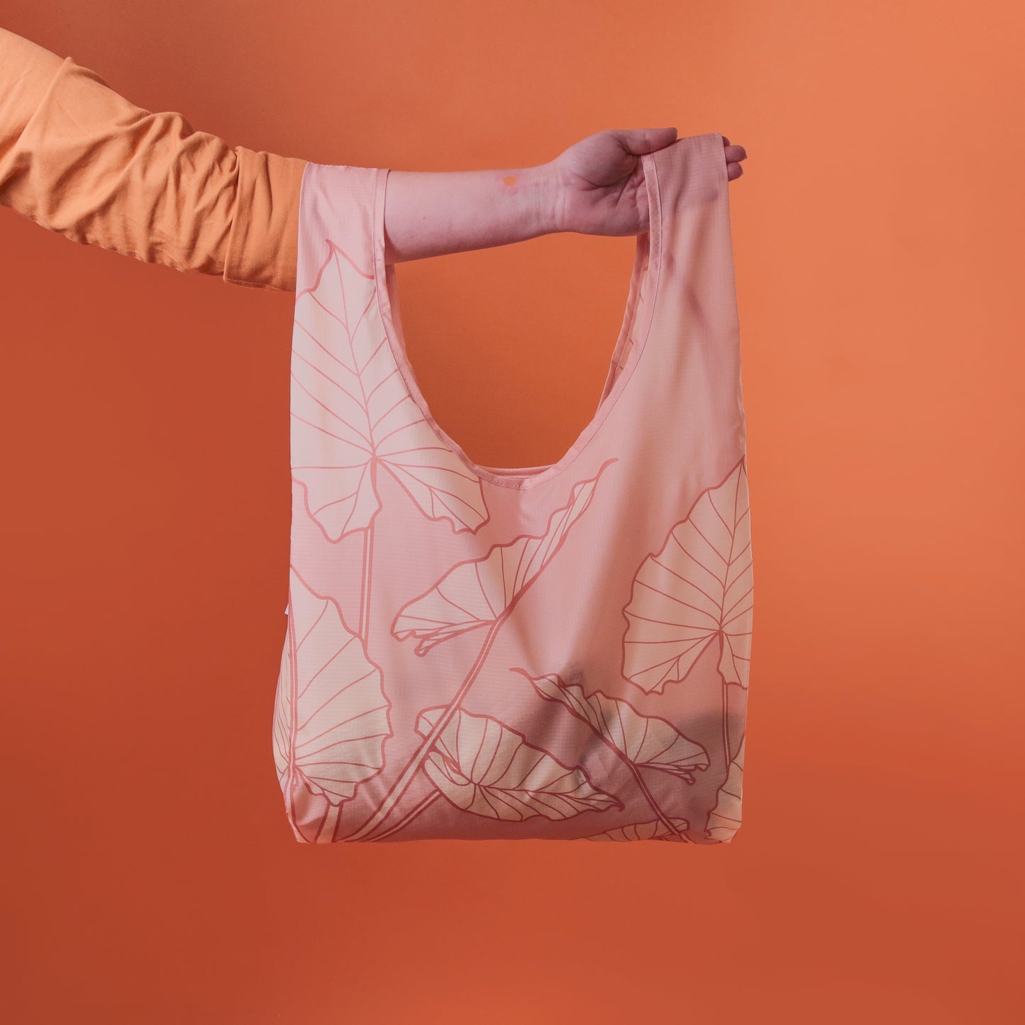 On a orange background is a light pink reusable tote bag made of nylon and detailed with line drawings of alocasia leaves.